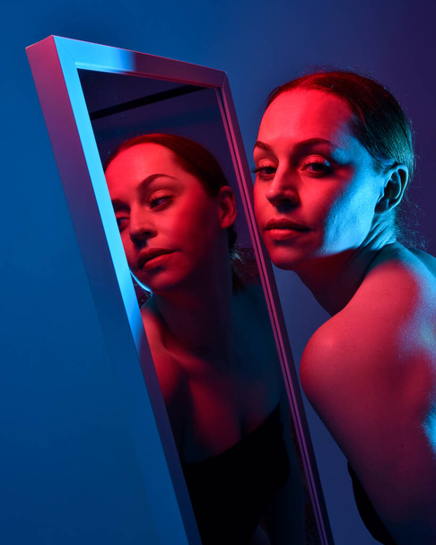 Κοντινό πλάνο πορτρέτο του όμορφου κοριτσιού κοιτάζοντας στον καθρέφτη, αυτο-αντανάκλαση με πολύχρωμο φωτισμό στούντιο. - Φωτογραφία, εικόνα