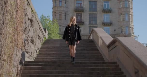 Adorable caucasian blonde girl in black jeans jacket going down on staircase on camera in European city center, springtime. High quality 4k video footage - Metraje, vídeo