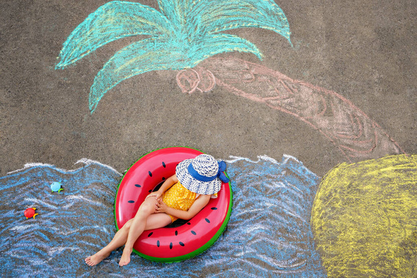 Счастливая маленькая дошкольница в купальнике на надувном кольце с морем, песком, ладонью, раскрашенной разноцветными мелом на асфальте. Симпатичный ребенок с веселым мелом. Лето, каникулы, лето. - Фото, изображение
