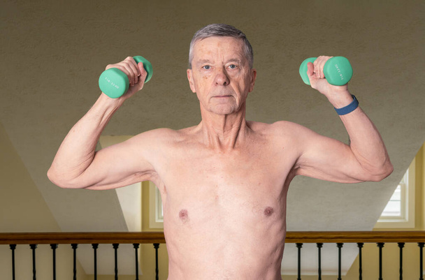 Shirtless senior man seen from front and exercising with dumbbells in home against railing - Fotoğraf, Görsel