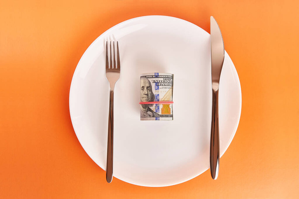 Белая тарелка с рулоном долларов, вилкой и ножом на оранжевом фоне. - Фото, изображение
