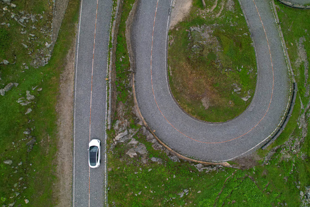 AERIAL, TOP DOWN: Brand new white car cruises along a scenic empty mountain pass road in Switzerland. Tourists on fun road trip cross the stunning San Gottardo Pass on a rainy summer afternoon. - Fotó, kép