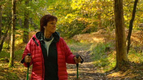 Female retiree unwinds by listening to an amusing podcast and exploring the colorful autumn forest. Elderly Caucasian woman listens to music while trekking down a forest trail. - Foto, Imagen