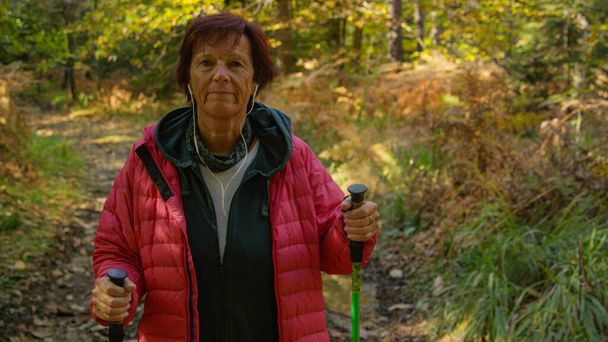 Smiling female retiree unwinds by listening to a podcast and exploring the colorful autumn forest. Elderly Caucasian woman listens to music while trekking down a forest trail. - Fotografie, Obrázek