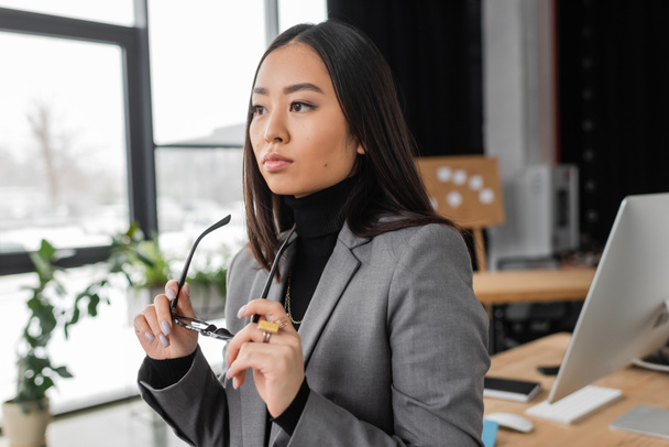pensive and young asian designer holding eyeglasses in office  - Фото, зображення