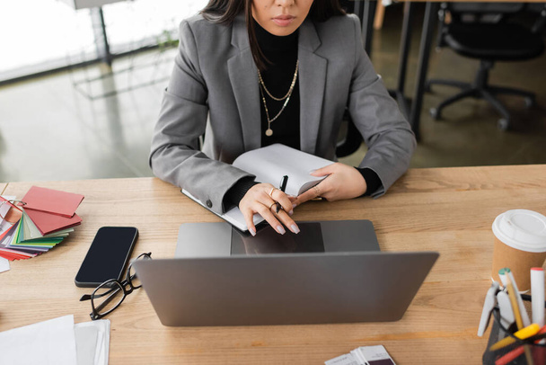 Обрезанный вид дизайнера интерьеров, держащего ноутбук рядом с устройствами и цветными образцами в офисе  - Фото, изображение
