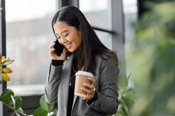 Positiivinen aasialainen suunnittelija puhuu matkapuhelimella ja pitää takeaway kahvia toimistossa  - Valokuva, kuva