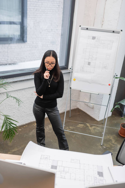 Високий кут зору спантеличеного азіатського дизайнера, що стоїть біля креслення і комп'ютерів в офісі
  - Фото, зображення