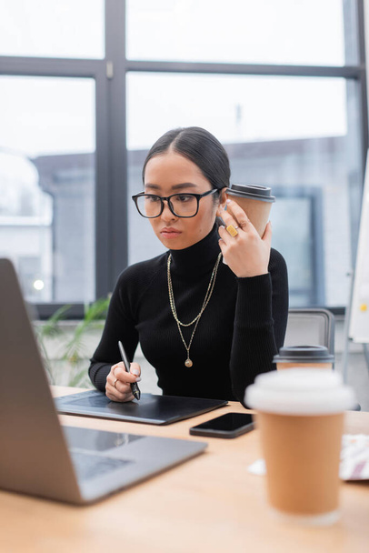 Азиатский дизайнер в очках держит бумажную чашку возле графического планшета и ноутбука в офисе  - Фото, изображение