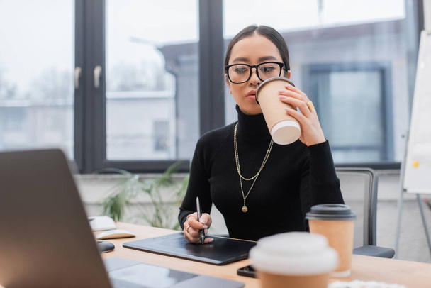 Asian designer holding coffee to go while using graphic tablet in office  - Photo, Image