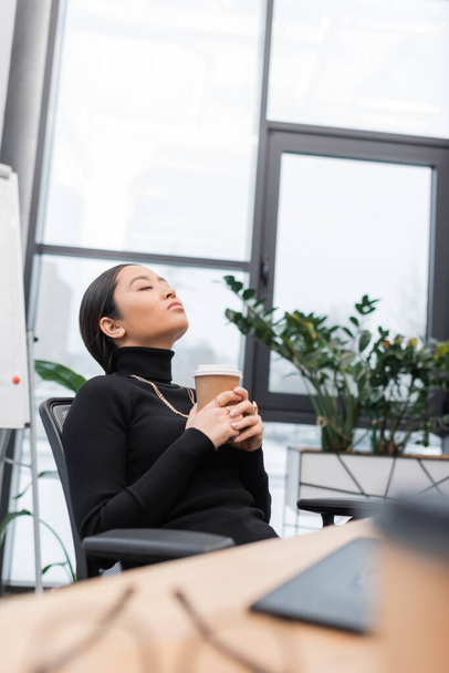 Asian designer holding coffee to go and relaxing near working table in office  - Photo, Image
