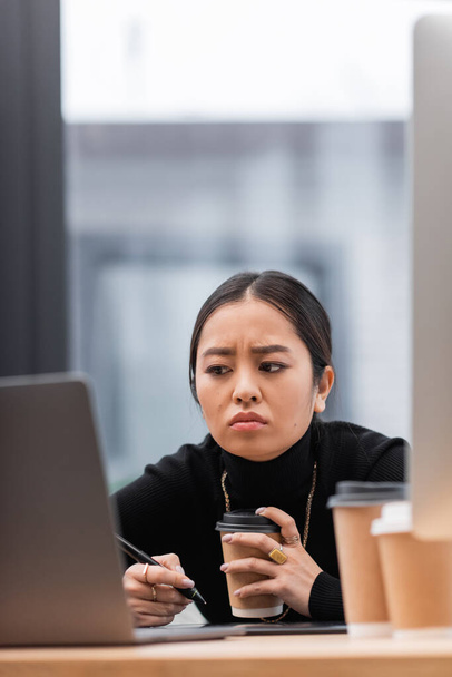 Displeased asian designer holding coffee near graphic tablet and computers in office  - Fotó, kép