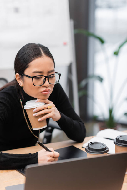 Набридливий азіатський дизайнер тримає каву, щоб піти і використовувати графічний планшет в офісі
  - Фото, зображення