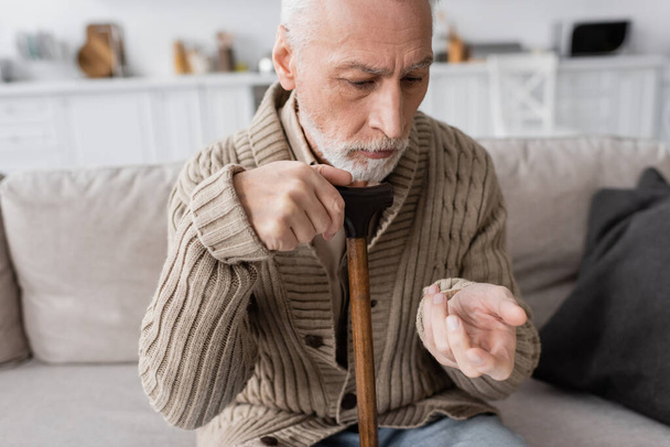 senior man in knitted cardigan sitting with walking cane and looking at trembling hand while suffering from parkinson disease - Photo, Image