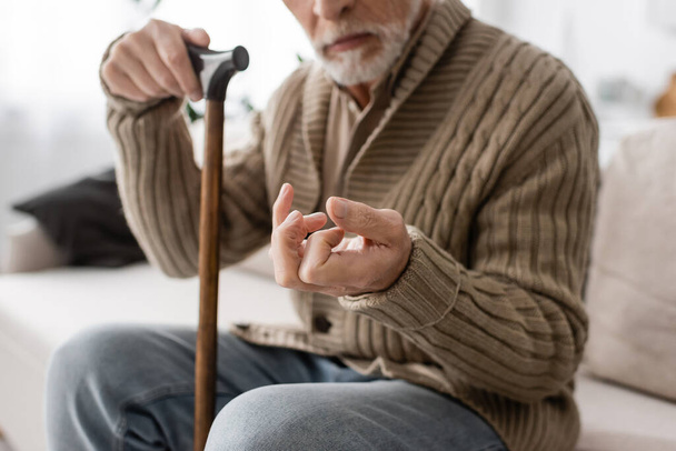 cropped view of senior man with parkinson disease holding walking cane while sitting on couch at home - Zdjęcie, obraz