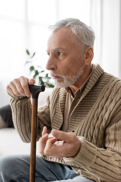 depressed man with parkinsonian syndrome sitting with walking cane and looking away at home - Foto, Imagen