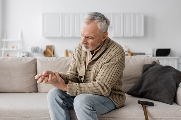 літній чоловік з синдромом Паркінсона страждає від тремтіння в руках, сидячи на дивані біля ходьби тростини
 - Фото, зображення