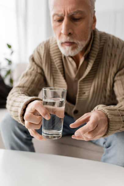 θολή άνθρωπος με σύνδρομο Parkinson και τρέμουλο στα χέρια κάθεται και κρατώντας ένα ποτήρι νερό στο σπίτι - Φωτογραφία, εικόνα