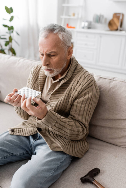 senior man with parkinson disease sitting on couch in knitted cardigan and holding pills in trembling hands - Fotó, kép