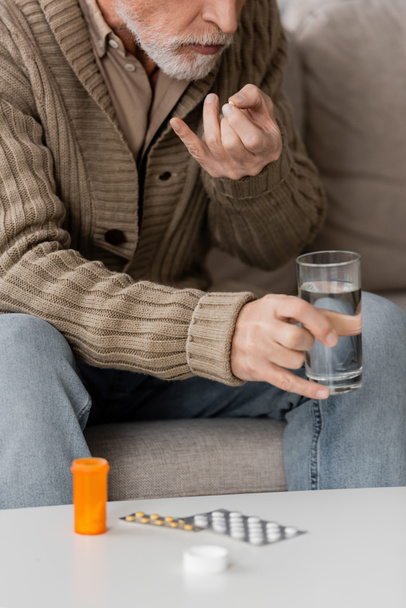 обрезанный вид человека с болезнью Паркинсона держа стакан воды возле таблеток на столе дома - Фото, изображение