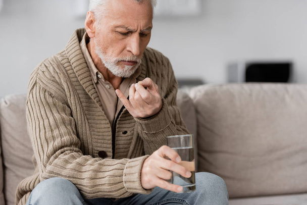 пожилой человек с синдромом Паркинсона сидит на диване и держит таблетки и стакан воды в дрожащих руках - Фото, изображение