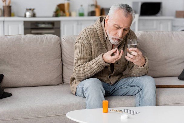 пожилой человек с синдромом Паркинсона держит таблетки и стакан воды, сидя на диване рядом с лекарствами на столе - Фото, изображение