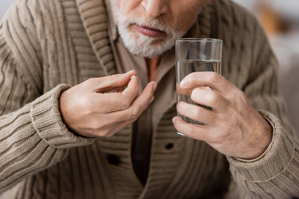 partial view of senior man with parkinson disease holding pill and glass of water in trembling hands - Foto, Imagen