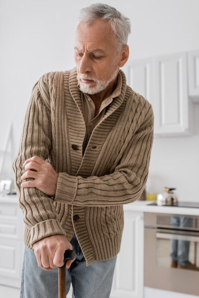 tensed man in knitted cardigan standing with walking cane in kitchen while suffering from parkinson disease - Photo, image