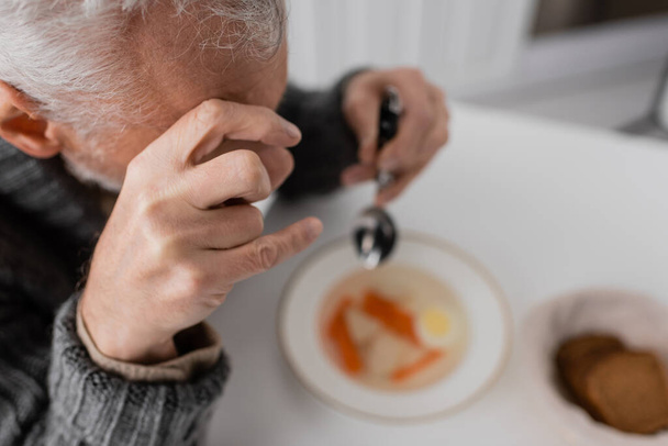 άποψη υψηλής γωνίας του ανθρώπου με σύνδρομο Parkinson και τρέμουν τα χέρια κρατώντας κουτάλι κοντά θολή σούπα στην κουζίνα - Φωτογραφία, εικόνα