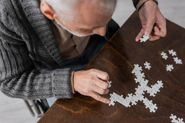 overhead view of senior man with parkinson disease and tremor in hands combining elements of jigsaw puzzle on table at home - Foto, imagen