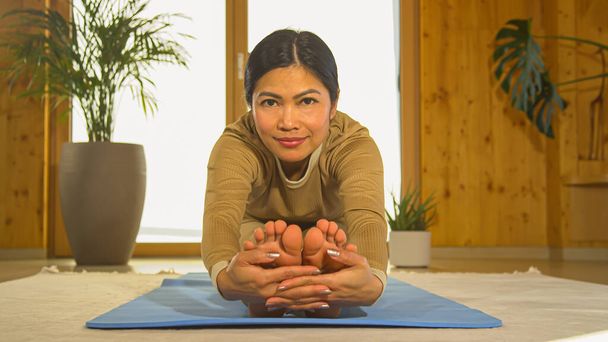 Young Filipino woman stretching her legs and working out at home. Asian female person doing seated forward fold yoga pose while looking at camera. Healthy leisure activity for vitality. - Foto, imagen