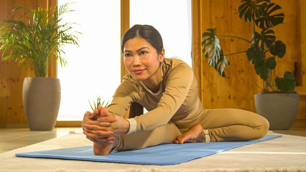 Young Asian woman performing seated head to knee yoga pose. Filipino female person doing body stretching on blue yoga mat. Yoga practice for healthy lifestyle, vitality and natural balance - Fotó, kép