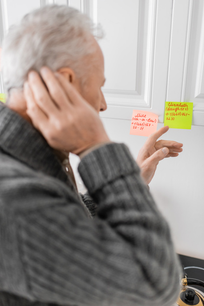 blurred man suffering from memory loss and pointing at sticky notes with names and phone numbers in kitchen - Photo, Image