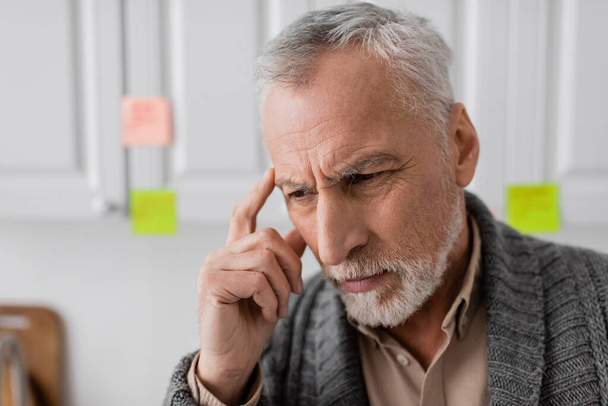thoughtful senior man with alzheimer disease touching head near blurred sticky notes in kitchen - Zdjęcie, obraz