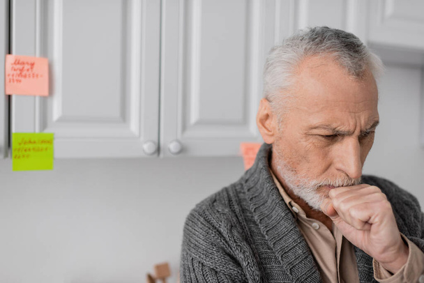 aged man with alzheimer syndrome holding fist near face while thinking near blurred sticky notes in kitchen - Photo, Image