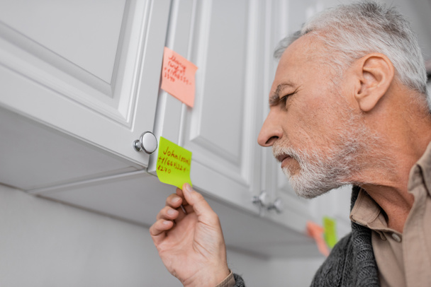 senior man with alzheimer syndrome looking at sticky note with name and phone number in kitchen - Foto, imagen