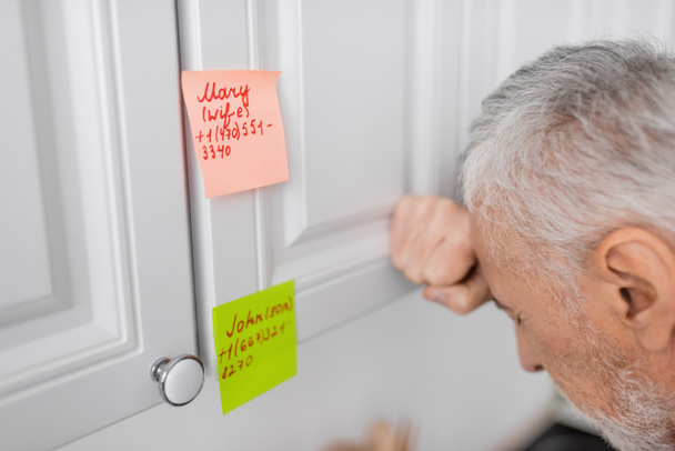 stressed man suffering from memory loss and standing near sticky notes with names and phone numbers in kitchen - Foto, imagen