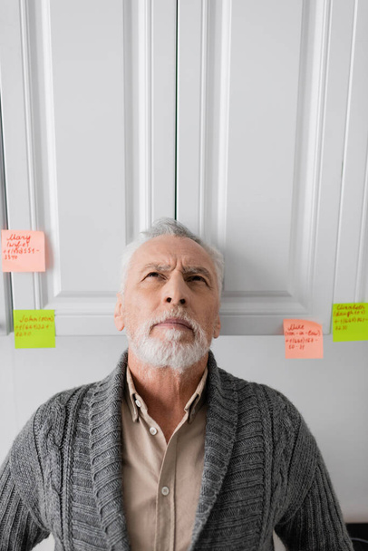 senior man suffering from memory loss and looking up while standing near sticky notes with names and phone numbers in kitchen - Fotografie, Obrázek