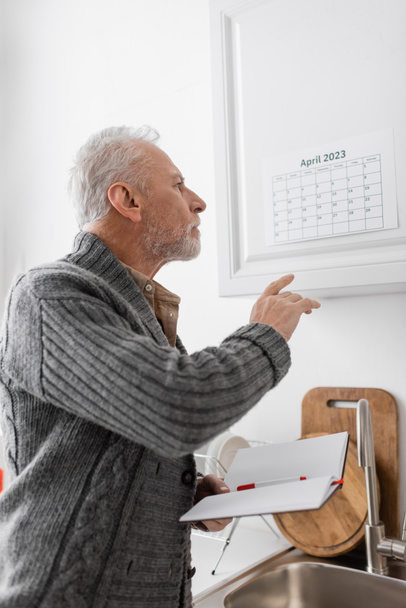 アルツハイマー病の老人がノートを持っていて台所でカレンダーを指しています - 写真・画像