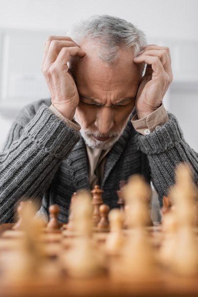 depressed man with alzheimer disease sitting with closed eyes and hands near head at chessboard on blurred foreground - Foto, imagen