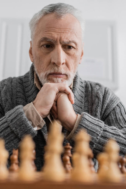 thoughtful senior man with alzheimer syndrome looking at blurred chessboard at home - Zdjęcie, obraz