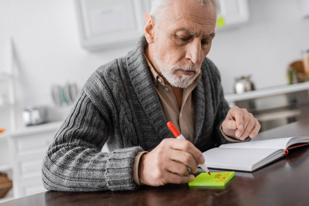 senior man with alzheimer syndrome writing on sticky note near blank notebook  - Zdjęcie, obraz