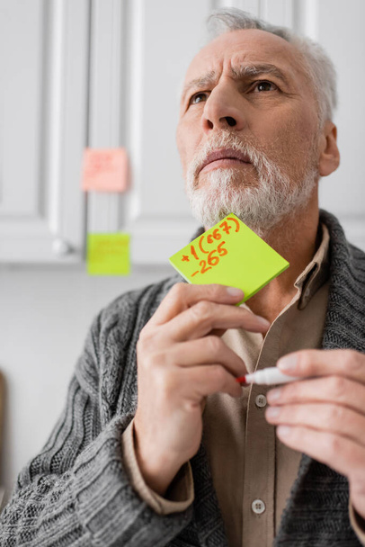 tense man suffering from memory loss while holding sticky notes with phone number and looking away in kitchen - Photo, Image