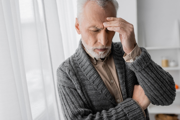 καταθλιπτικός ηλικιωμένος άνδρας με σύνδρομο Αλτσχάιμερ στέκεται με κλειστά μάτια και αγγίζει το μέτωπο στο σπίτι - Φωτογραφία, εικόνα