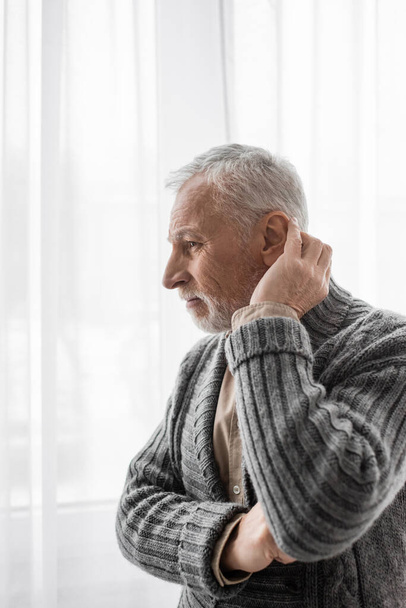 старший и вдумчивый мужчина в вязаном кардигане, глядя в окно, страдающий синдромом Альцгеймера - Фото, изображение