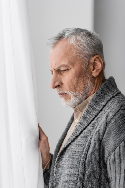 side view of depressed senior man with alzheimer diseased looking away near window at home - Foto, Imagen