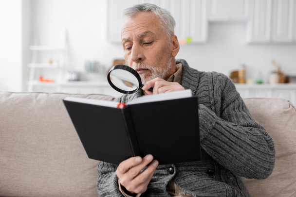senior man with azheimers disease looking in notebook through magnifier while sitting on couch at home - Fotó, kép