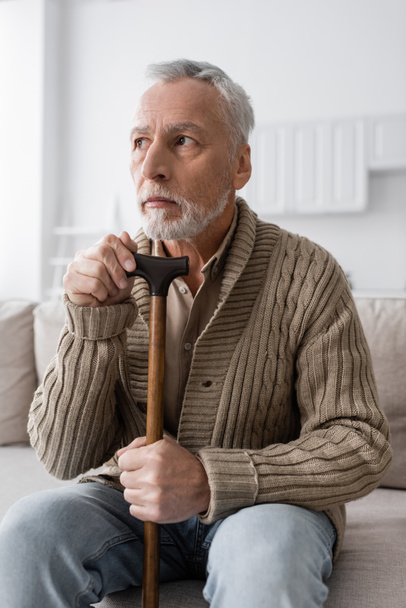 senior man with alzheimer disease looking away while sitting with walking cane at home - Fotó, kép