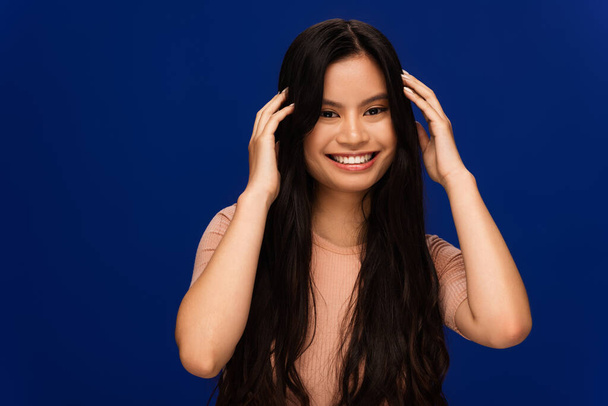 Cheerful asian woman touching long hair isolated on blue  - Fotografie, Obrázek
