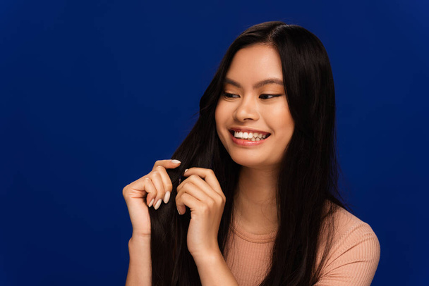 Cheerful asian woman looking at hair isolated on blue  - Photo, Image
